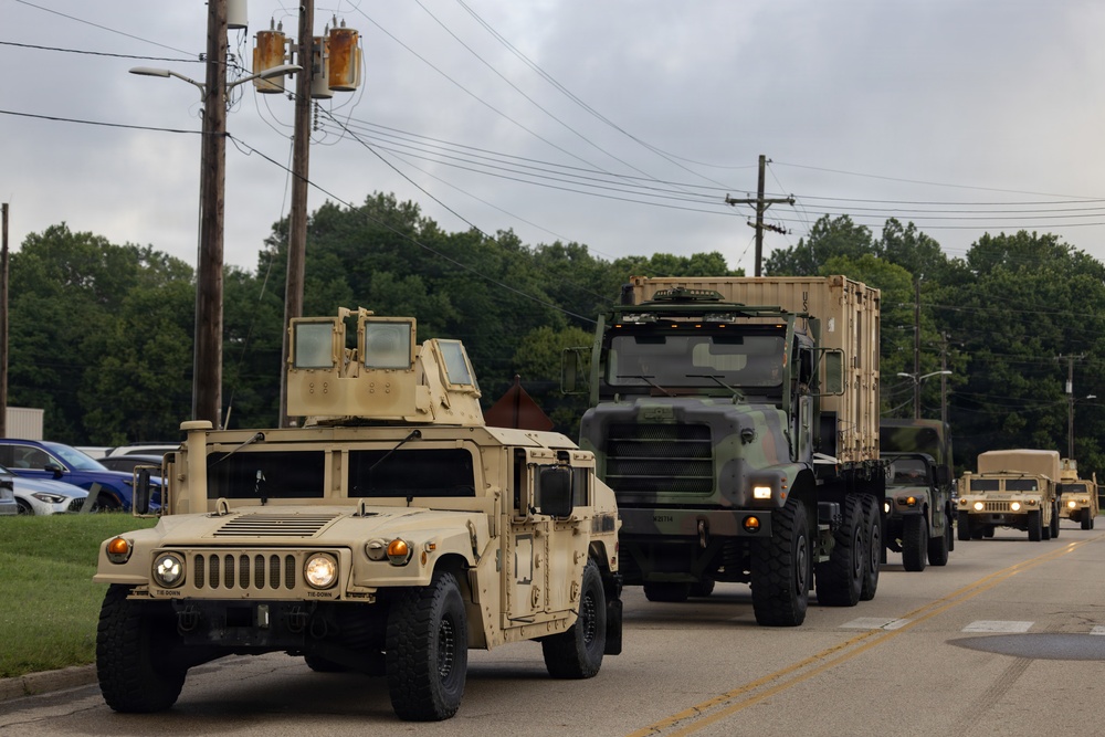 A military convoy is traveling along a two-lane road, led by a tan/desert-colored HMMWV (Humvee) followed by a camouflage-painted heavy military truck hauling a large cargo container. Additional military vehicles, including at least one more Humvee, are visible further back in the convoy. The setting appears to be a suburban or semi-rural area with trees, utility poles, and a parking lot visible in the background.