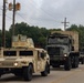 A military convoy is traveling along a two-lane road, led by a tan/desert-colored HMMWV (Humvee) followed by a camouflage-painted heavy military truck hauling a large cargo container. Additional military vehicles, including at least one more Humvee, are visible further back in the convoy. The setting appears to be a suburban or semi-rural area with trees, utility poles, and a parking lot visible in the background.