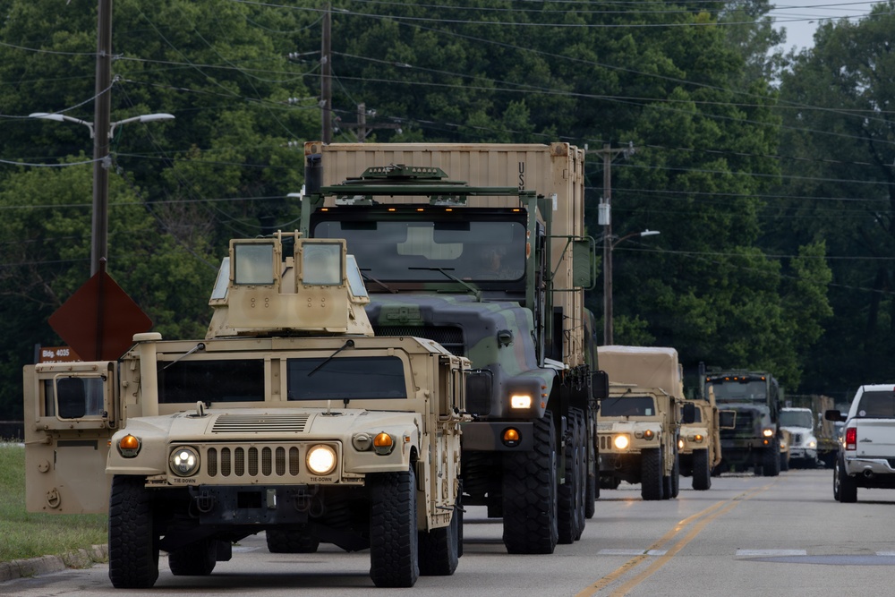 A military convoy of U.S. Army vehicles is traveling along a paved road, led by a tan/desert-colored HMMWV (Humvee) followed by a large military cargo truck carrying a shipping container marked 'US'. The convoy extends into the background with multiple additional military vehicles visible, suggesting a significant troop or equipment movement operation.