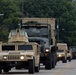 A military convoy of U.S. Army vehicles is traveling along a paved road, led by a tan/desert-colored HMMWV (Humvee) followed by a large military cargo truck carrying a shipping container marked 'US'. The convoy extends into the background with multiple additional military vehicles visible, suggesting a significant troop or equipment movement operation.