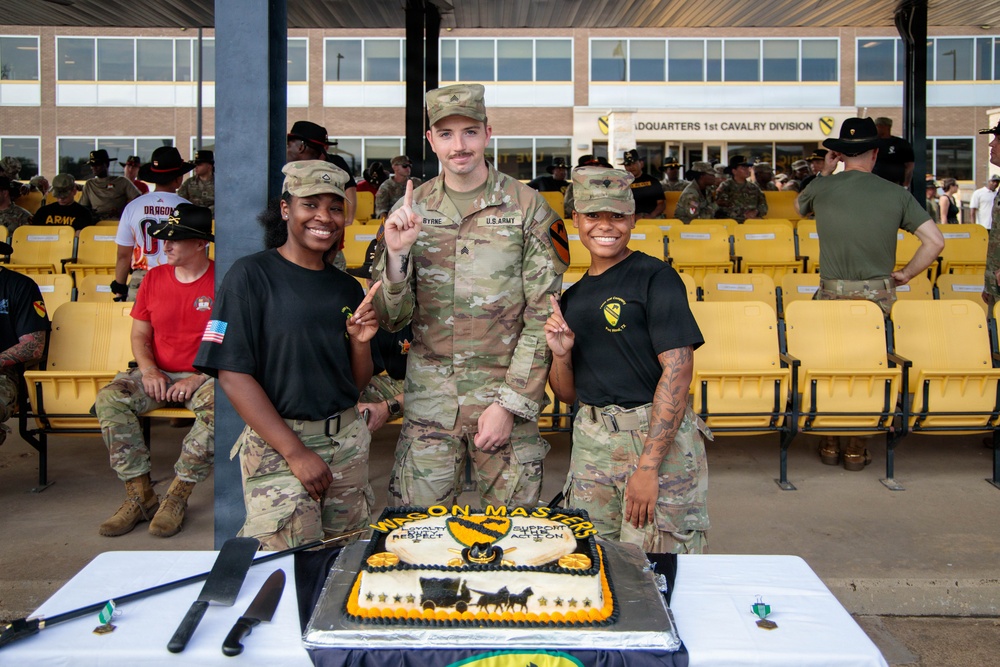 Three U.S. Army soldiers pose for a photo in front of a decorated cake at what appears to be a military ceremony or celebration at the 1st Cavalry Division Headquarters. The soldier in the center is in full Army Combat Uniform (ACU) and is holding up one finger, flanked by two female soldiers in black Army PT-style shirts. A large decorated cake reading 'Wilson Masters' with 1st Cavalry Division insignia is displayed on the table in front of them.