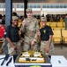 Three U.S. Army soldiers pose for a photo in front of a decorated cake at what appears to be a military ceremony or celebration at the 1st Cavalry Division Headquarters. The soldier in the center is in full Army Combat Uniform (ACU) and is holding up one finger, flanked by two female soldiers in black Army PT-style shirts. A large decorated cake reading 'Wilson Masters' with 1st Cavalry Division insignia is displayed on the table in front of them.