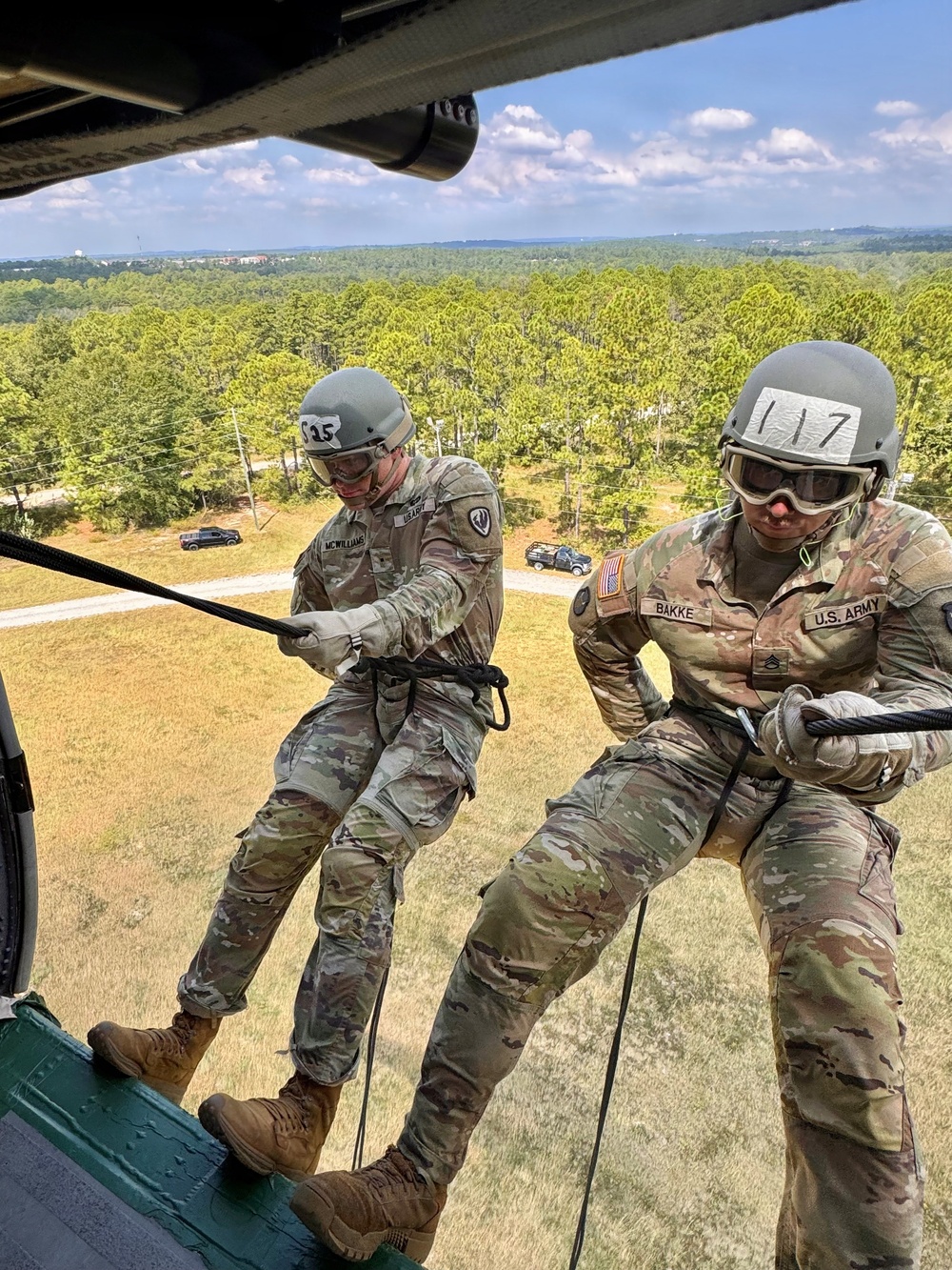 Two U.S. Army soldiers are rappelling from a military helicopter during a training exercise. Both soldiers are wearing combat uniforms, helmets with identification numbers (S25 and 117), and protective goggles while gripping rappel ropes. The image is taken from inside the helicopter looking outward, with a forested landscape and road visible below.