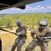 Two U.S. Army soldiers are rappelling from a military helicopter during a training exercise. Both soldiers are wearing combat uniforms, helmets with identification numbers (S25 and 117), and protective goggles while gripping rappel ropes. The image is taken from inside the helicopter looking outward, with a forested landscape and road visible below.