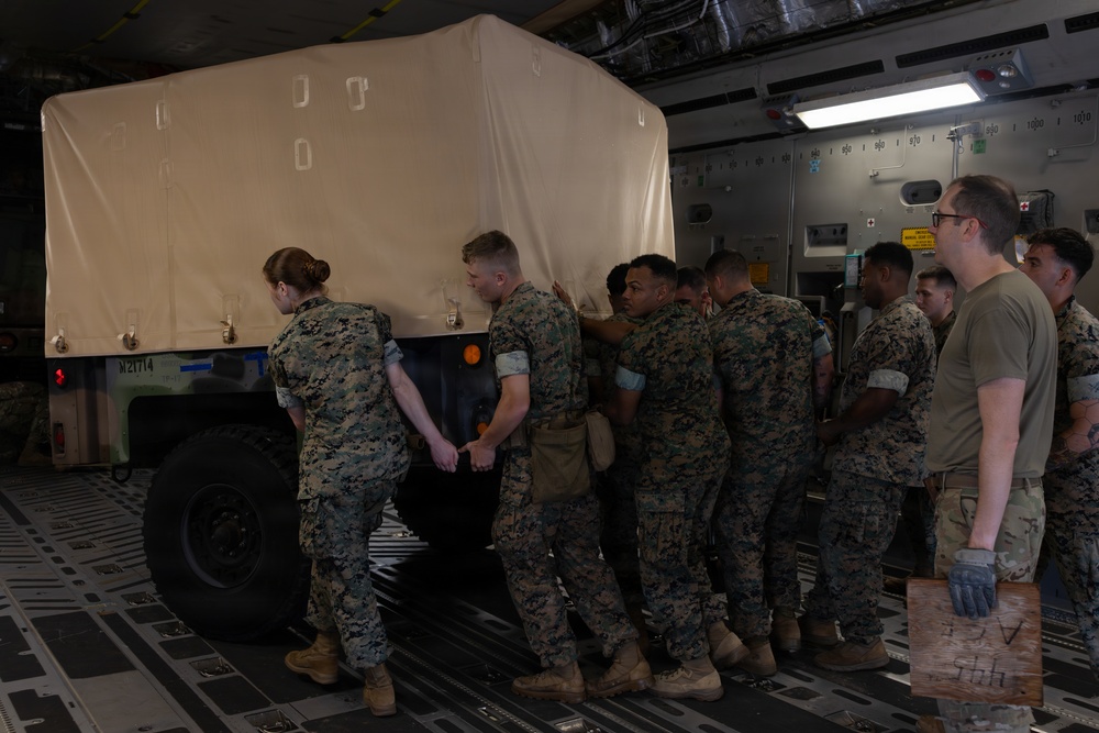 Military personnel in camouflage uniforms are loading or positioning a canvas-covered military vehicle inside what appears to be a large military transport aircraft. A group of approximately 8-10 service members are pushing or guiding the vehicle into position on the aircraft's cargo floor. The setting is the interior of a large cargo aircraft, identifiable by the distinctive ribbed flooring and interior wall panels.
