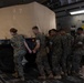 Military personnel in camouflage uniforms are loading or positioning a canvas-covered military vehicle inside what appears to be a large military transport aircraft. A group of approximately 8-10 service members are pushing or guiding the vehicle into position on the aircraft's cargo floor. The setting is the interior of a large cargo aircraft, identifiable by the distinctive ribbed flooring and interior wall panels.