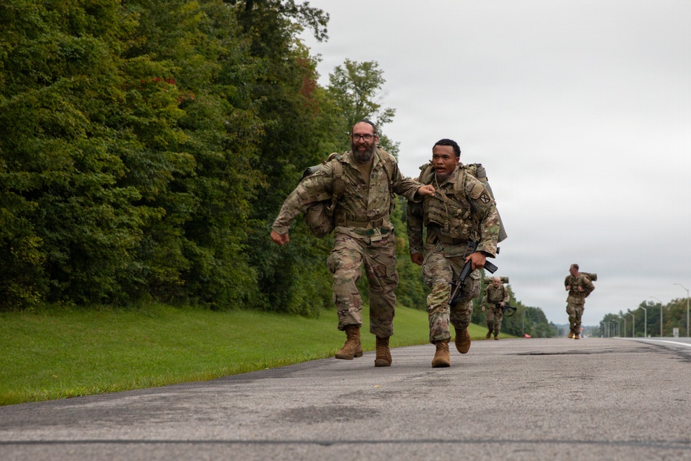 Two soldiers in U.S. Army OCP (Operational Camouflage Pattern) uniforms are running together on a paved road, with one appearing to support or encourage the other. Both are carrying full rucksacks and tactical gear, consistent with a ruck march or physical fitness event. Additional soldiers are visible in the background completing the same activity.