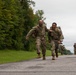 Two soldiers in U.S. Army OCP (Operational Camouflage Pattern) uniforms are running together on a paved road, with one appearing to support or encourage the other. Both are carrying full rucksacks and tactical gear, consistent with a ruck march or physical fitness event. Additional soldiers are visible in the background completing the same activity.