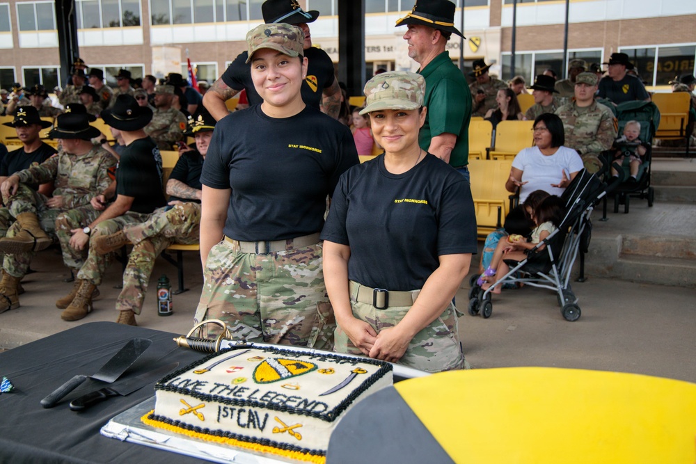 Two female soldiers in U.S. Army uniforms wearing 'Stay Ironhorse' black t-shirts and camouflage pants pose behind a ceremonial cake at what appears to be a 1st Cavalry Division event. The cake is decorated with the text 'Live The Legend - 1st CAV' along with unit insignia in black, gold, and white. A large military formation or gathering is visible in the background, with soldiers in various uniforms including campaign hats.
