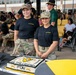 Two female soldiers in U.S. Army uniforms wearing 'Stay Ironhorse' black t-shirts and camouflage pants pose behind a ceremonial cake at what appears to be a 1st Cavalry Division event. The cake is decorated with the text 'Live The Legend - 1st CAV' along with unit insignia in black, gold, and white. A large military formation or gathering is visible in the background, with soldiers in various uniforms including campaign hats.