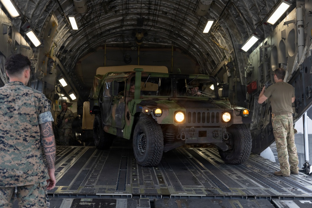 A military HMMWV (Humvee) is being loaded or unloaded from the cargo bay of a large military transport aircraft, likely a C-17 Globemaster III. Several military personnel in different uniforms are present, guiding and overseeing the vehicle movement. The headlights of the Humvee are illuminated, suggesting active operation.