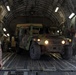 A military HMMWV (Humvee) is being loaded or unloaded from the cargo bay of a large military transport aircraft, likely a C-17 Globemaster III. Several military personnel in different uniforms are present, guiding and overseeing the vehicle movement. The headlights of the Humvee are illuminated, suggesting active operation.
