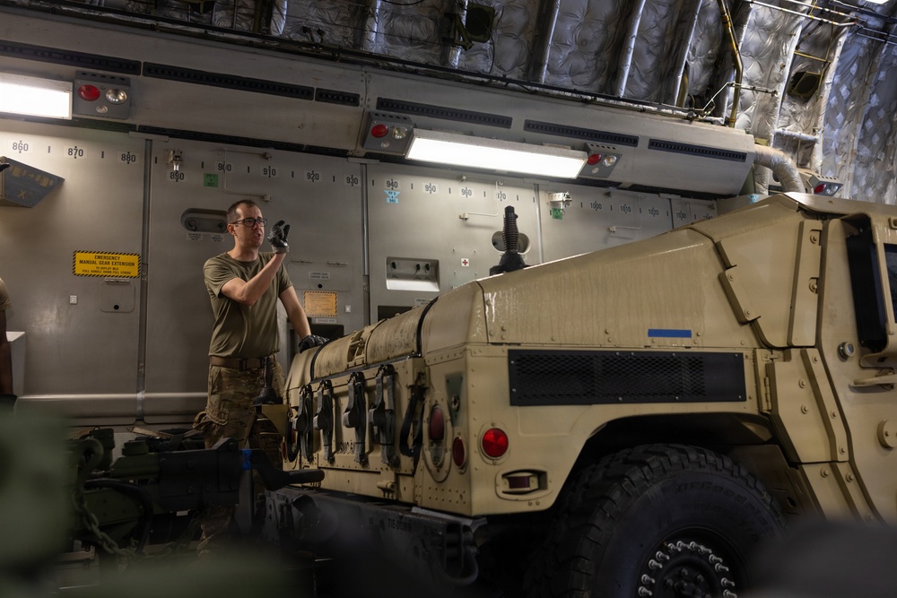 A military service member in combat uniform is working inside what appears to be the cargo hold of a large military transport aircraft, likely a C-17 Globemaster or similar. A tan/desert-colored HMMWV (Humvee) military vehicle is being loaded or secured inside the aircraft. The service member appears to be signaling or directing the vehicle positioning while wearing gloves.