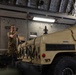 A military service member in combat uniform is working inside what appears to be the cargo hold of a large military transport aircraft, likely a C-17 Globemaster or similar. A tan/desert-colored HMMWV (Humvee) military vehicle is being loaded or secured inside the aircraft. The service member appears to be signaling or directing the vehicle positioning while wearing gloves.