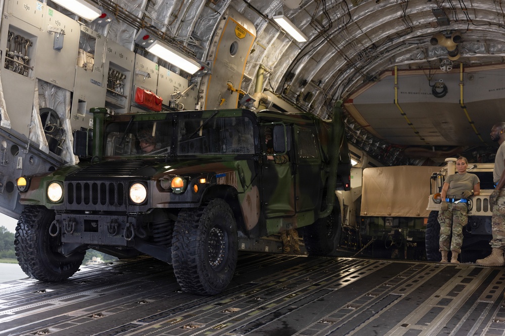 A military HMMWV (Humvee) in olive drab camouflage is being loaded or unloaded from the cargo bay of a large military transport aircraft, likely a C-17 Globemaster III. The vehicle's headlights are illuminated and personnel in military uniforms are guiding the operation. At least one additional tan/desert-colored military vehicle is visible further inside the cargo hold.
