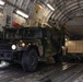 A military HMMWV (Humvee) in olive drab camouflage is being loaded or unloaded from the cargo bay of a large military transport aircraft, likely a C-17 Globemaster III. The vehicle's headlights are illuminated and personnel in military uniforms are guiding the operation. At least one additional tan/desert-colored military vehicle is visible further inside the cargo hold.