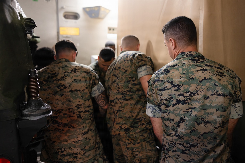 A group of military personnel wearing MARPAT (Marine Pattern) digital camouflage uniforms are gathered together in a confined space, with their backs to the camera. They appear to be huddled around something, possibly reviewing equipment or a document. The setting appears to be inside a military vehicle or facility.