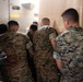 A group of military personnel wearing MARPAT (Marine Pattern) digital camouflage uniforms are gathered together in a confined space, with their backs to the camera. They appear to be huddled around something, possibly reviewing equipment or a document. The setting appears to be inside a military vehicle or facility.