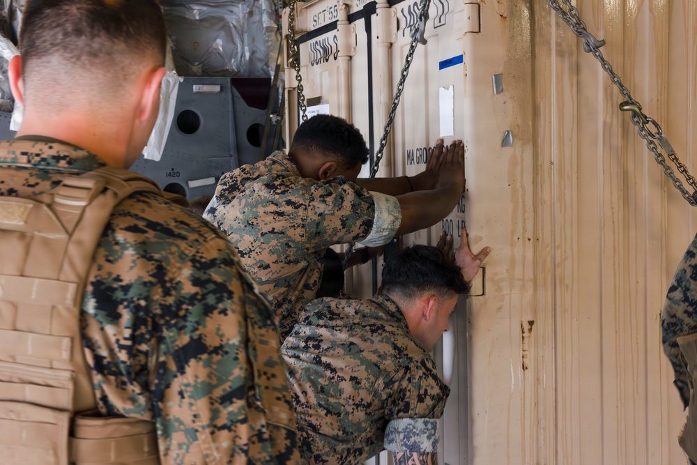 Several military personnel in MARPAT (Marine pattern) camouflage uniforms are working with a large tan shipping/storage container secured with chains. Two soldiers are actively pushing or examining the container door while a third observes from the foreground. The setting appears to be inside a cargo aircraft or military transport vehicle.