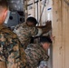 Several military personnel in MARPAT (Marine pattern) camouflage uniforms are working with a large tan shipping/storage container secured with chains. Two soldiers are actively pushing or examining the container door while a third observes from the foreground. The setting appears to be inside a cargo aircraft or military transport vehicle.