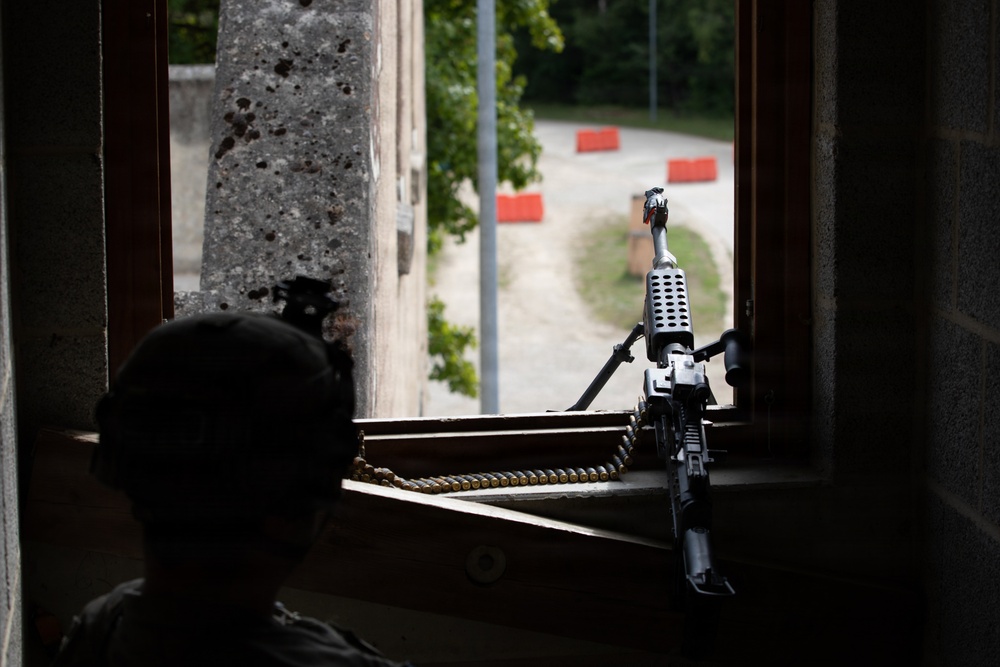 A silhouetted figure wearing a helmet is positioned at a window with a belt-fed machine gun resting on the windowsill, aimed outward toward a road with orange barriers. The building wall shows visible pockmarks consistent with bullet damage. The scene appears to depict a defensive firing position, likely during a military training exercise or operational deployment.