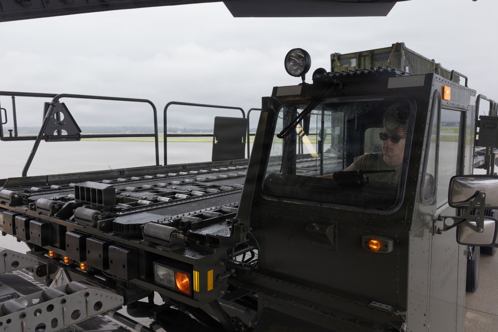 A military ground support vehicle, likely an aircraft cargo loader or munitions handling vehicle, is being operated on what appears to be a military airfield tarmac. A single operator wearing sunglasses and a military-style olive drab shirt is visible through the cab window. The overcast weather conditions and wet surfaces suggest recent or ongoing rainfall.