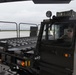 A military ground support vehicle, likely an aircraft cargo loader or munitions handling vehicle, is being operated on what appears to be a military airfield tarmac. A single operator wearing sunglasses and a military-style olive drab shirt is visible through the cab window. The overcast weather conditions and wet surfaces suggest recent or ongoing rainfall.