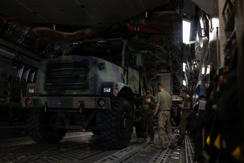 Military personnel are inside the cargo hold of a large military transport aircraft, likely a C-17 Globemaster III, with a camouflaged tactical military truck loaded aboard. Three soldiers in camouflage uniforms appear to be reviewing documents or a clipboard near the vehicle. The scene depicts a military airlift or air transport loading/inspection operation.