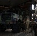 Military personnel are inside the cargo hold of a large military transport aircraft, likely a C-17 Globemaster III, with a camouflaged tactical military truck loaded aboard. Three soldiers in camouflage uniforms appear to be reviewing documents or a clipboard near the vehicle. The scene depicts a military airlift or air transport loading/inspection operation.