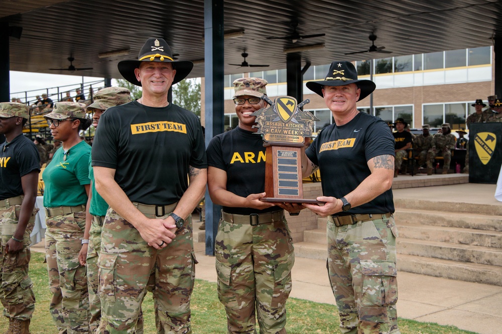 Three U.S. Army soldiers pose together holding a trophy at what appears to be an outdoor military ceremony. The trophy is labeled 'Assault Kitchen Cook-Off Competition' for the 1st Cavalry Division. Two of the individuals wear distinctive black cowboy-style hats with gold insignia and 'First Team' black t-shirts, consistent with 1st Cavalry Division leadership attire.