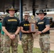 Three U.S. Army soldiers pose together holding a trophy at what appears to be an outdoor military ceremony. The trophy is labeled 'Assault Kitchen Cook-Off Competition' for the 1st Cavalry Division. Two of the individuals wear distinctive black cowboy-style hats with gold insignia and 'First Team' black t-shirts, consistent with 1st Cavalry Division leadership attire.