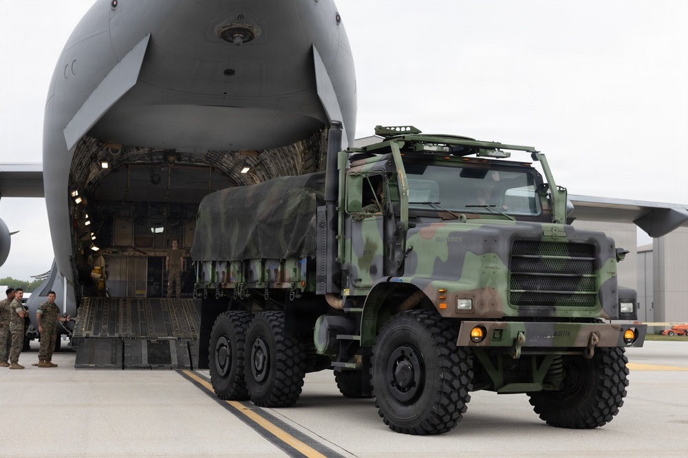 A military cargo operation is underway at an airfield, with a camouflage-painted heavy tactical truck being offloaded from a large military transport aircraft. The truck, consistent with a Medium Tactical Vehicle Replacement (MTVR) or similar 6x6 military cargo truck, is driving down the aircraft's rear loading ramp. Several military personnel in uniform are visible supervising the operation on the tarmac.