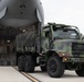 A military cargo operation is underway at an airfield, with a camouflage-painted heavy tactical truck being offloaded from a large military transport aircraft. The truck, consistent with a Medium Tactical Vehicle Replacement (MTVR) or similar 6x6 military cargo truck, is driving down the aircraft's rear loading ramp. Several military personnel in uniform are visible supervising the operation on the tarmac.