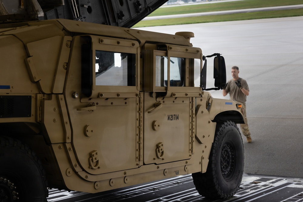 A tan/desert-colored military HMMWV (Humvee) bearing the identification number M21714 is being loaded or unloaded from a military transport aircraft via a rear cargo ramp. A soldier in tan/coyote brown uniform is visible in the background, appearing to guide or marshal the vehicle. The setting is an airfield tarmac, with the aircraft's rear cargo door open overhead.