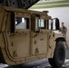 A tan/desert-colored military HMMWV (Humvee) bearing the identification number M21714 is being loaded or unloaded from a military transport aircraft via a rear cargo ramp. A soldier in tan/coyote brown uniform is visible in the background, appearing to guide or marshal the vehicle. The setting is an airfield tarmac, with the aircraft's rear cargo door open overhead.