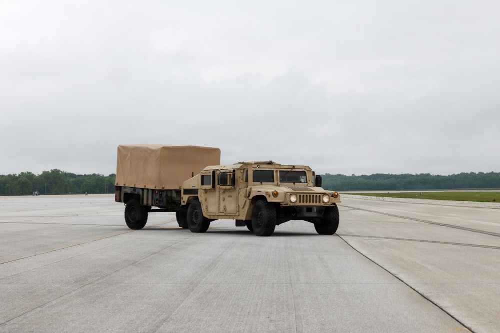 A tan/desert-colored military HMMWV (Humvee) is traveling across a large concrete tarmac or airfield, towing a covered flatbed trailer of matching color. The vehicle appears to be in operational condition with headlights on. The setting appears to be a military airfield or installation, with tree lines visible in the background under an overcast sky.
