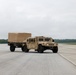 A tan/desert-colored military HMMWV (Humvee) is traveling across a large concrete tarmac or airfield, towing a covered flatbed trailer of matching color. The vehicle appears to be in operational condition with headlights on. The setting appears to be a military airfield or installation, with tree lines visible in the background under an overcast sky.