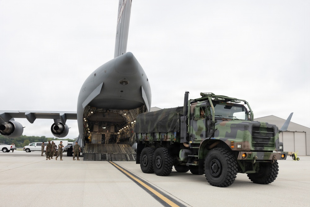 A military loading operation is underway at an airfield, where a camouflage-painted heavy military truck is being loaded into or unloaded from a large military transport aircraft. The aircraft's nose is raised, revealing the front cargo ramp and illuminated interior. Several military personnel are visible on the tarmac observing or directing the operation.