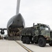 A military loading operation is underway at an airfield, where a camouflage-painted heavy military truck is being loaded into or unloaded from a large military transport aircraft. The aircraft's nose is raised, revealing the front cargo ramp and illuminated interior. Several military personnel are visible on the tarmac observing or directing the operation.