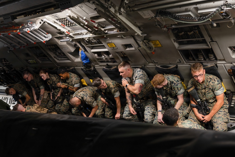 A group of military personnel in camouflage uniforms are seated inside the cargo hold of a large military transport aircraft. They appear to be preparing or waiting during a transport mission, with several individuals handling rifles and gear. The interior shows the characteristic ribbed metal structure and mechanical components of a large military cargo aircraft, likely a C-17 Globemaster or similar.