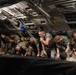 A group of military personnel in camouflage uniforms are seated inside the cargo hold of a large military transport aircraft. They appear to be preparing or waiting during a transport mission, with several individuals handling rifles and gear. The interior shows the characteristic ribbed metal structure and mechanical components of a large military cargo aircraft, likely a C-17 Globemaster or similar.