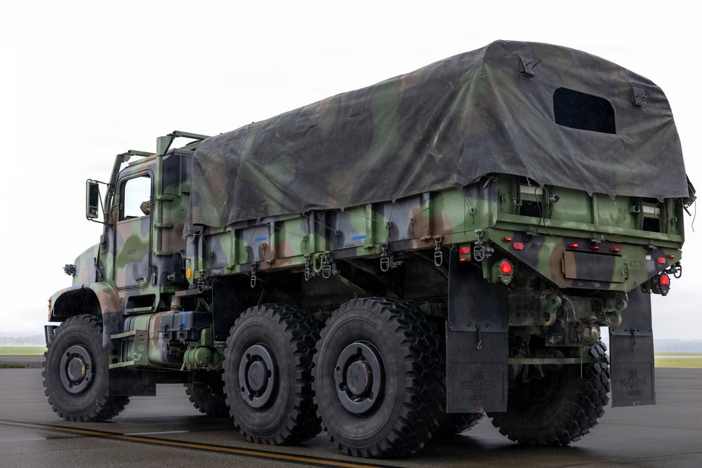 A military cargo truck in woodland camouflage pattern (green, brown, black) is parked on what appears to be a wet tarmac or airfield surface. The vehicle is a heavy-duty multi-axle truck, consistent with a US Army M939 or similar FMTV/LMTV series, fitted with a canvas cargo cover over the bed. The overcast sky and wet pavement suggest rainy or overcast weather conditions.