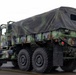 A military cargo truck in woodland camouflage pattern (green, brown, black) is parked on what appears to be a wet tarmac or airfield surface. The vehicle is a heavy-duty multi-axle truck, consistent with a US Army M939 or similar FMTV/LMTV series, fitted with a canvas cargo cover over the bed. The overcast sky and wet pavement suggest rainy or overcast weather conditions.