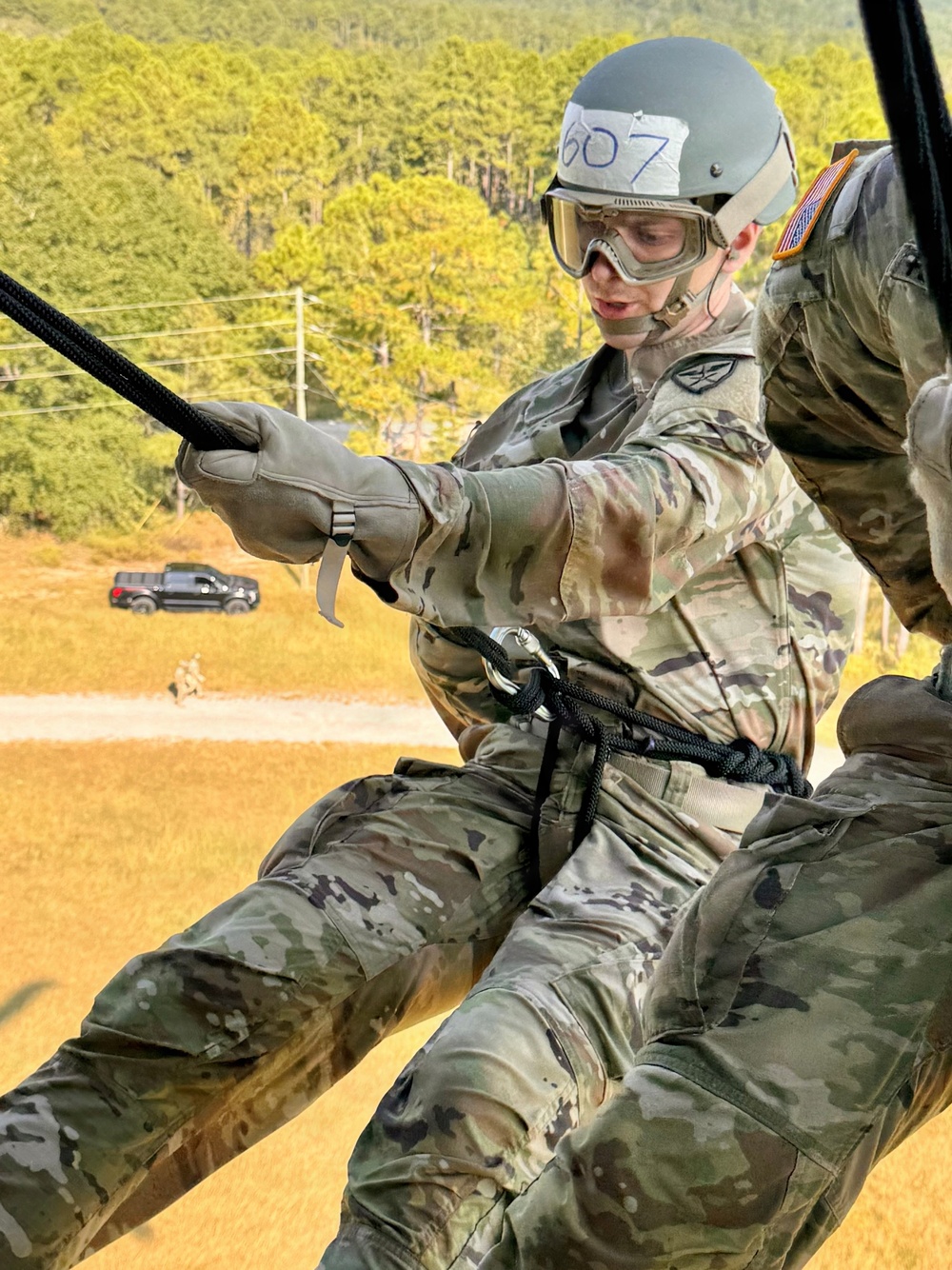 A U.S. Army soldier wearing OCP (Operational Camouflage Pattern) uniform is performing a rappelling or fast-rope descent exercise from an elevated position, likely a helicopter or training tower. The soldier is equipped with a helmet marked '607', protective goggles, gloves, and a rappelling harness with a black rope and carabiner. The background shows a grassy training area with pine trees, a dirt road, a dark pickup truck, and at least one other person visible on the ground.