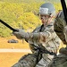 A U.S. Army soldier wearing OCP (Operational Camouflage Pattern) uniform is performing a rappelling or fast-rope descent exercise from an elevated position, likely a helicopter or training tower. The soldier is equipped with a helmet marked '607', protective goggles, gloves, and a rappelling harness with a black rope and carabiner. The background shows a grassy training area with pine trees, a dirt road, a dark pickup truck, and at least one other person visible on the ground.