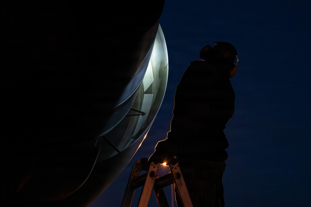 A person is standing on a ladder at night, inspecting or working on a large curved metallic structure, likely a satellite dish or radar antenna. The scene is dramatically lit with a bright light source illuminating the interior of the dish, casting the worker in silhouette against a deep blue night sky. A small warm light is also visible near the base of the ladder.