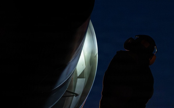 A person is standing on a ladder at night, inspecting or working on a large curved metallic structure, likely a satellite dish or radar antenna. The scene is dramatically lit with a bright light source illuminating the interior of the dish, casting the worker in silhouette against a deep blue night sky. A small warm light is also visible near the base of the ladder.