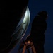 A person is standing on a ladder at night, inspecting or working on a large curved metallic structure, likely a satellite dish or radar antenna. The scene is dramatically lit with a bright light source illuminating the interior of the dish, casting the worker in silhouette against a deep blue night sky. A small warm light is also visible near the base of the ladder.