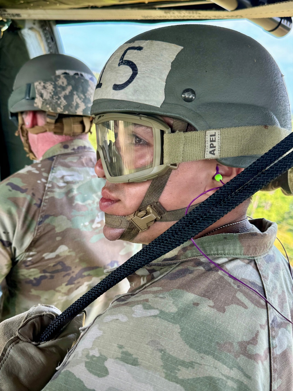 Two military personnel are seated inside what appears to be a military helicopter or aircraft. The foreground soldier wears a ballistic helmet marked with the number '5', APEL-labeled protective goggles, ear protection, and holds a dark rope or static line. Both individuals are dressed in MultiCam/OCP camouflage uniforms.