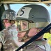Two military personnel are seated inside what appears to be a military helicopter or aircraft. The foreground soldier wears a ballistic helmet marked with the number '5', APEL-labeled protective goggles, ear protection, and holds a dark rope or static line. Both individuals are dressed in MultiCam/OCP camouflage uniforms.